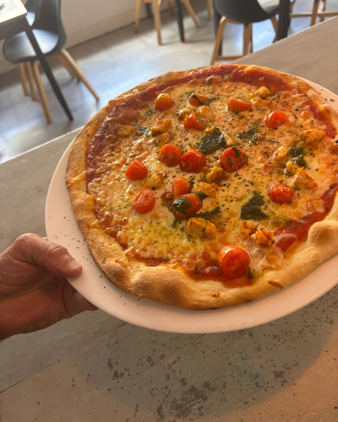 Mano sujetando un plato blanco con una pizza de tomate cherry, pollo y pesto